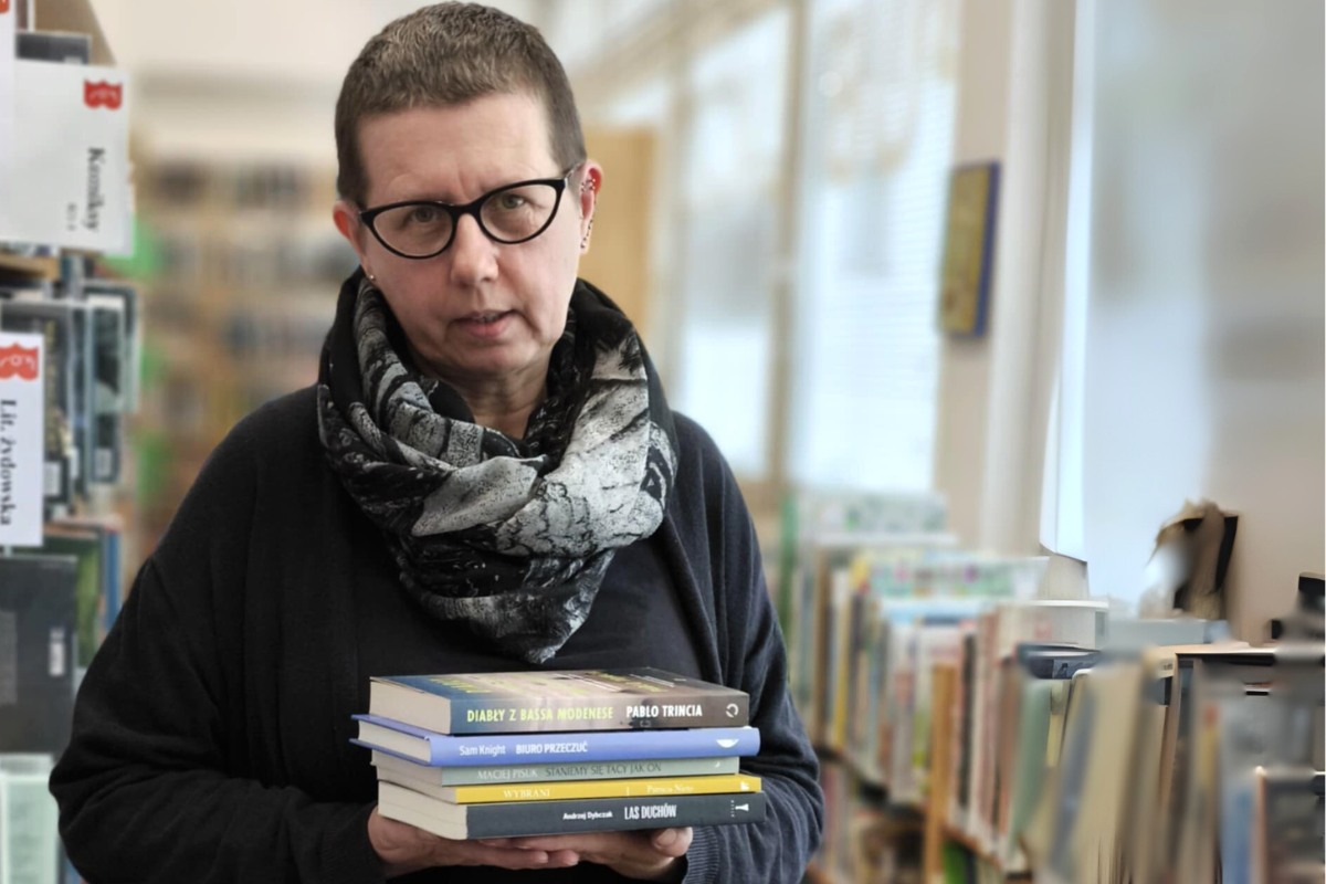 Kobieta w czarnej bluzie i szalu trzyma stos książek, stojąc w bibliotece z regałami w tle.